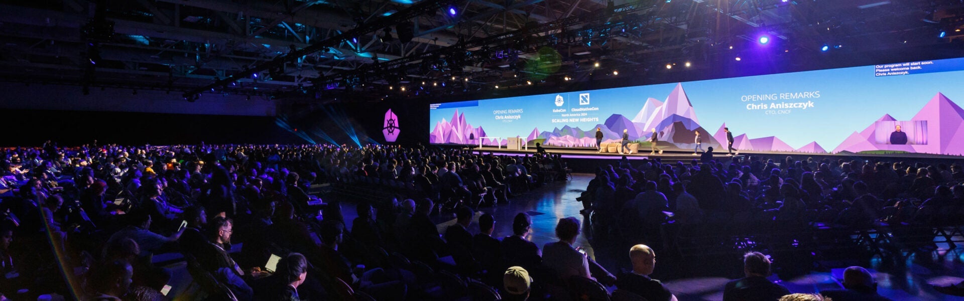 A wide angle view from the back of a keynote speaker room showing a large audience and 5 speakers walking onto the stage.